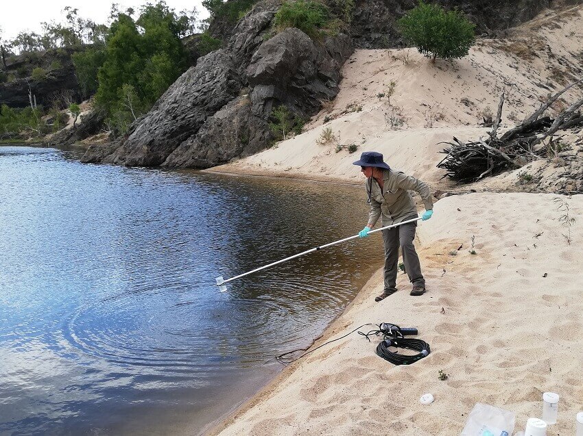 Person wearing gloves and using a pole to dip a hand-sized test tube into black river water