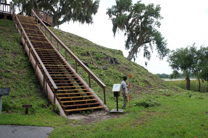A very large midden containing oysters covered in grass with wooden stairs built to the top and a person standing in front