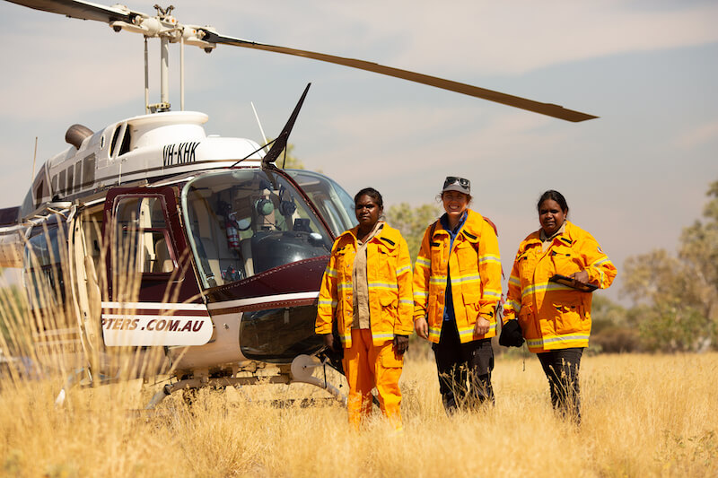 Three people in high vis suits stand outside a helicopter surrounded by yellow grass