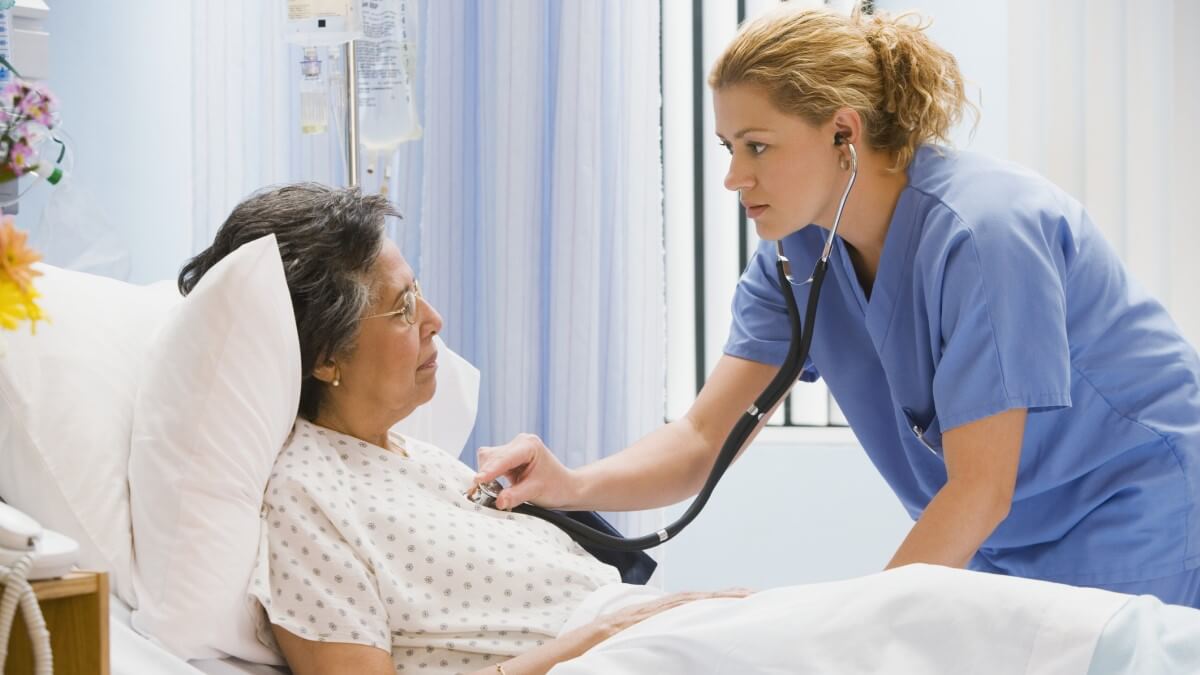 woman using a stethoscope on an older patient in a hospital bed