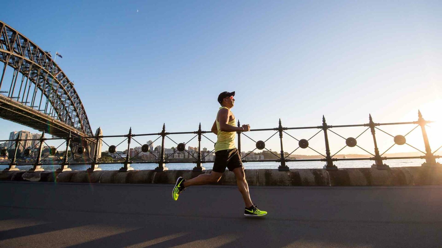 A man in a singlet and bright yellow shoes runs along Sydney harbour foreshroe with the Sydney harbour bridge in the backgrond