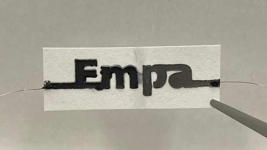 Photo of the paper battery. It is a piece of paper with 'empa' written on it in thick black and grey text. The e and the a are both connected to wires at either side of the paper. The paper between the m and the p is visibly damp.