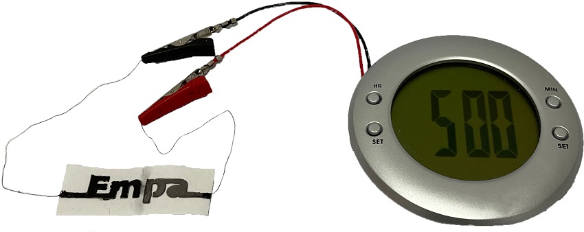 Paper battery connected to a small round clock via red and black alligator clips connected to the battery wires