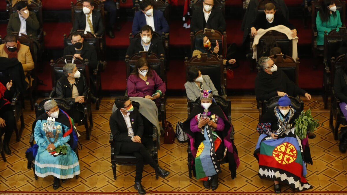 Chilean people sitting in chairs, some wearing traditional dress and some wearing suits, all wearing masks, in the hall of a grand room