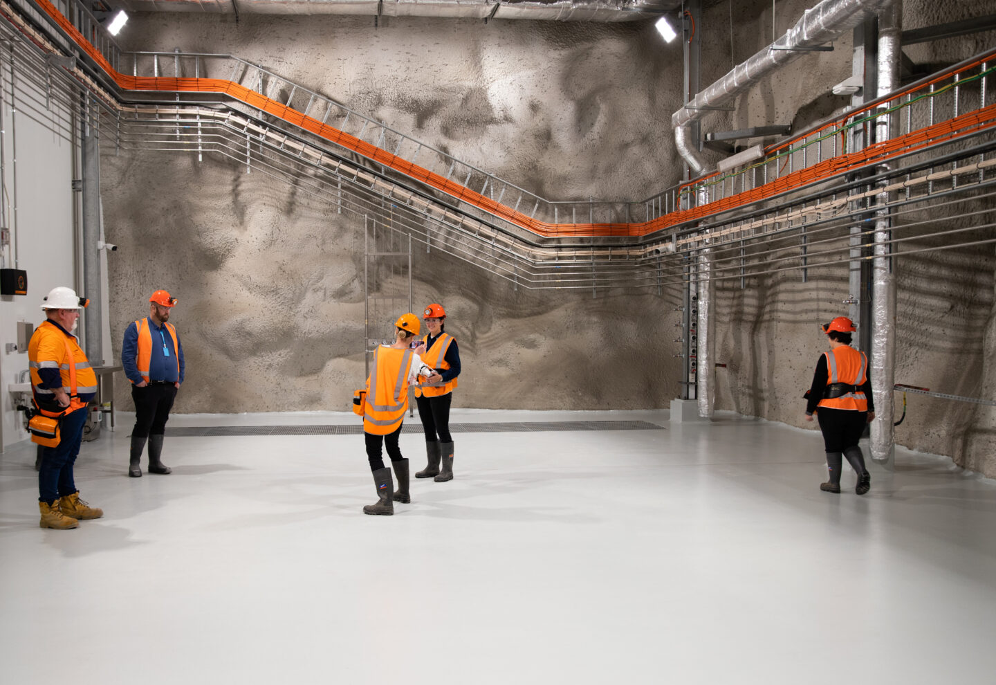 A cave like space with piped on the walls and six people in high vis and orange helmets walking around