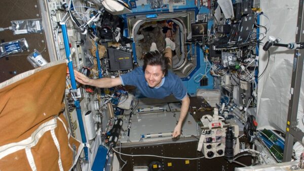 A man in blue floating in the middle of the frame. There's technology and wires all around him. He's grinning to the camera.