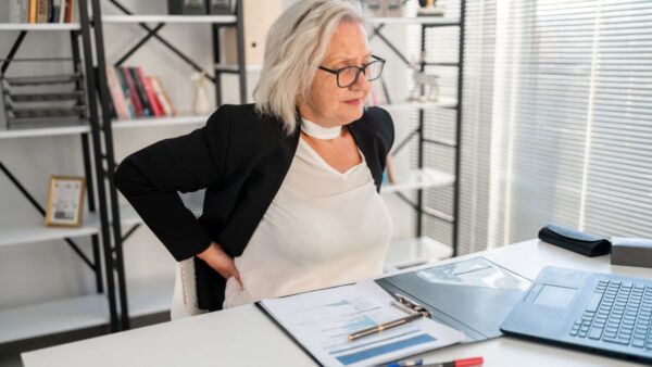Photograph of an elderly businesswoman holding her lower back in pain while seated at her desk at work