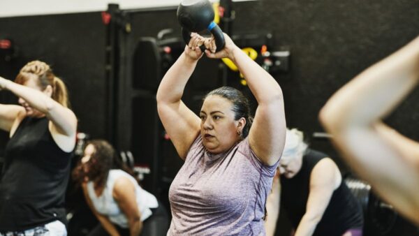 A mid-aged woman in a purple t-shirt swings a kettle bell above her head in a group gym class