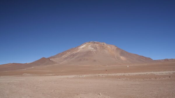 A photograph of a brown desert, with a mountaing rising above surrounded by clear blue skies