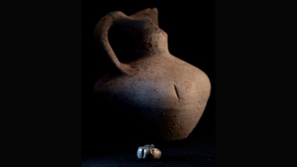 Photograph of a dented gold earring in front of a small clay pot. The objects are placed in front of a black background and surface.