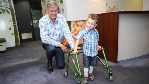 Photograph of an older man crouching next to a young child who is using a green walking frame to assist him to stand